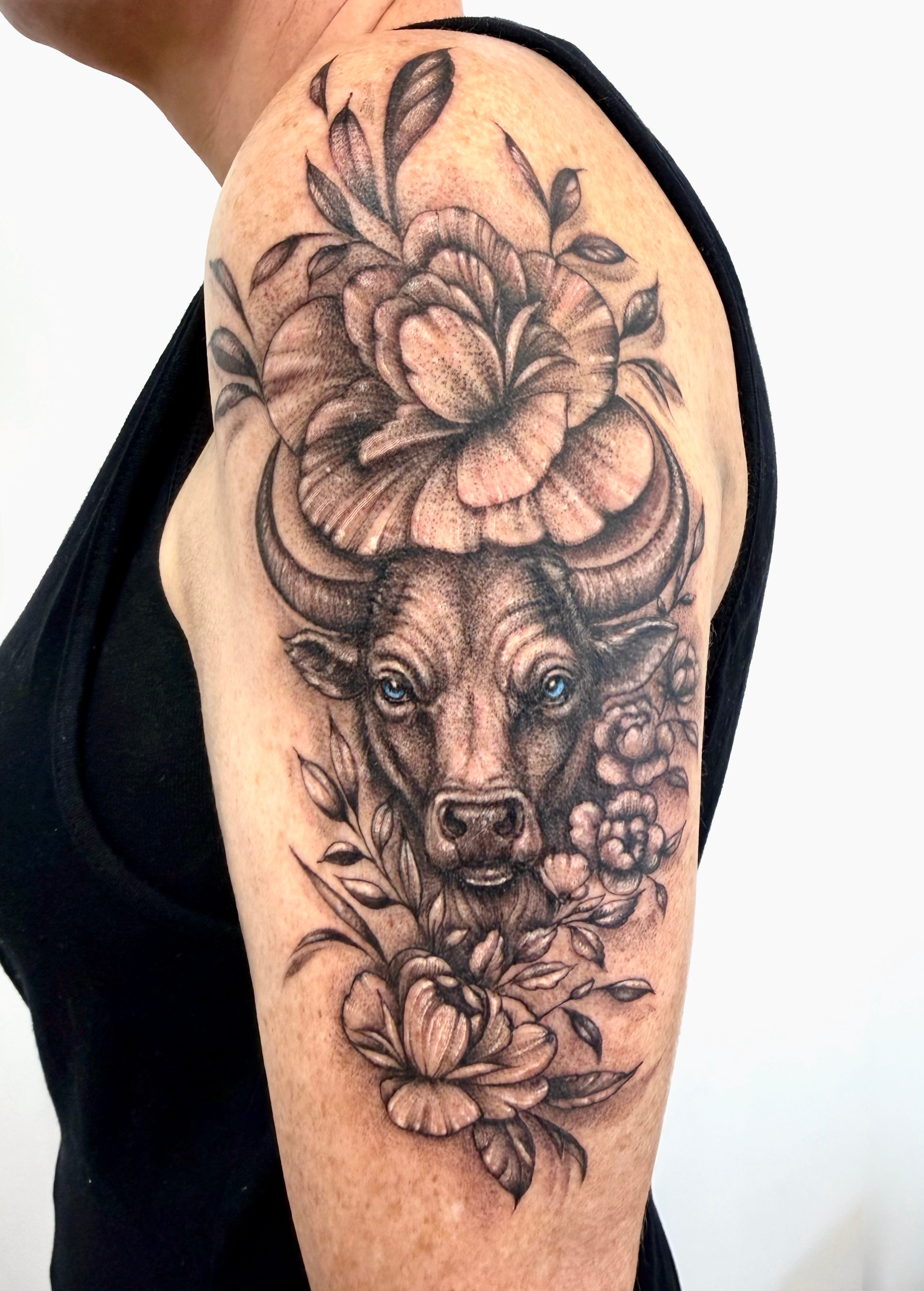 Bull with blue eyes surrounded by black and grey flowers and leaves, tattooed on the shoulder and upper arm