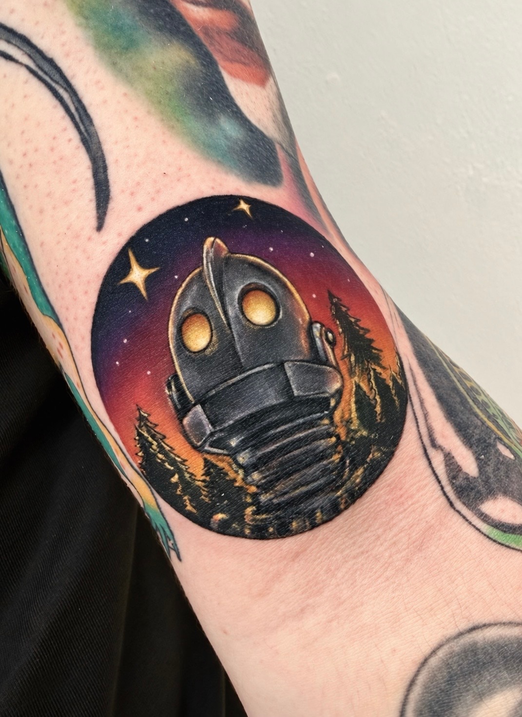 Iron Giant colour forearm cover-up tattoo with glowing eyes, pine trees and a starry orange and purple sky