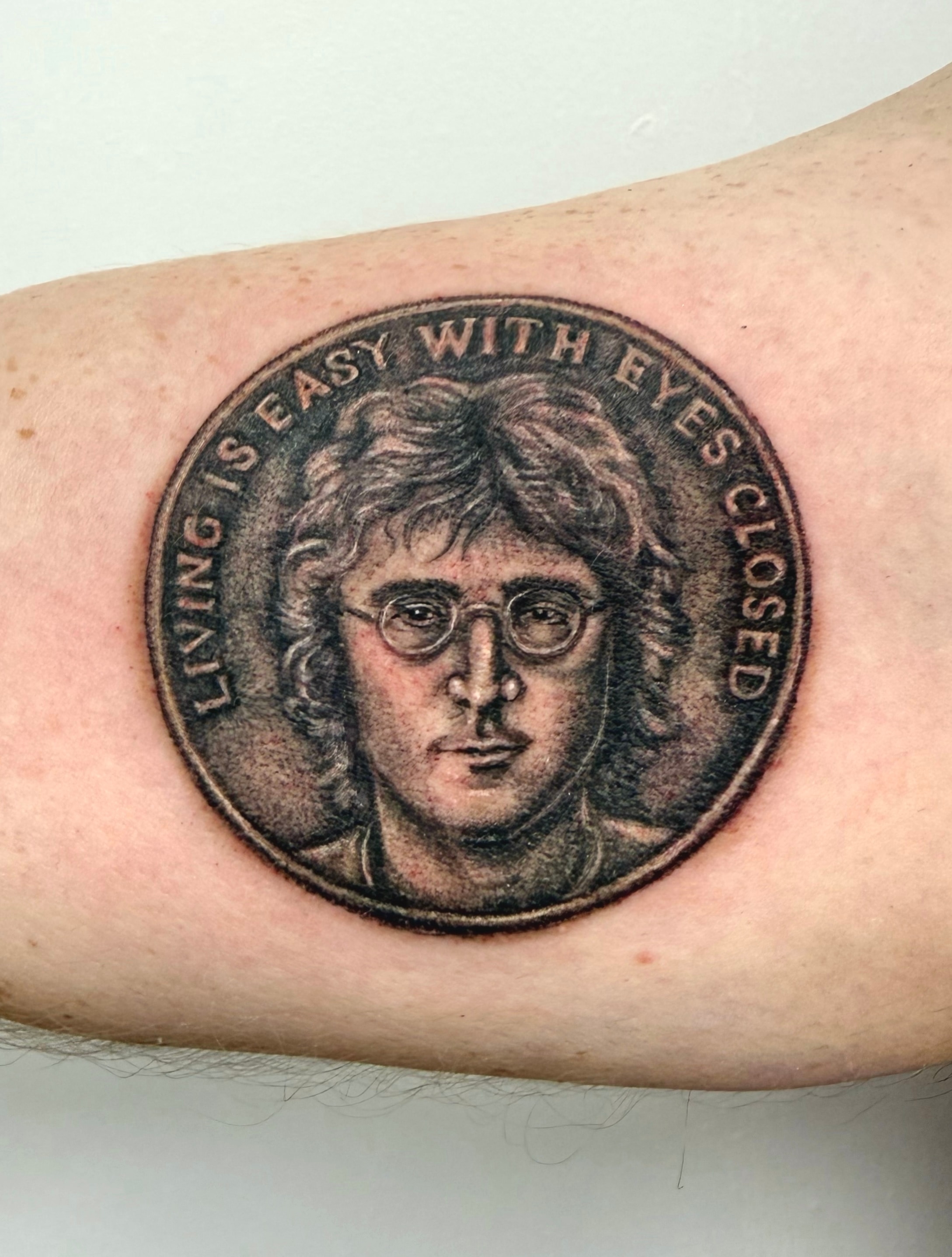 Colour portrait tattoo of John Lennon in round glasses, framed by a coin-shaped border and a song-lyric inscription on the upper arm
