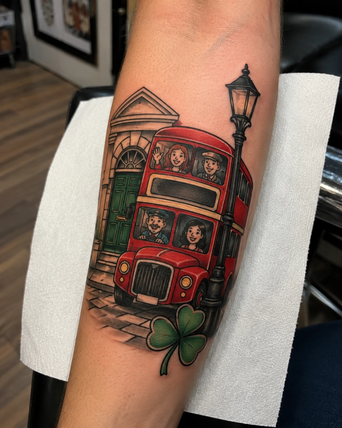 Red London bus tattoo with passengers and shamrock on forearm
