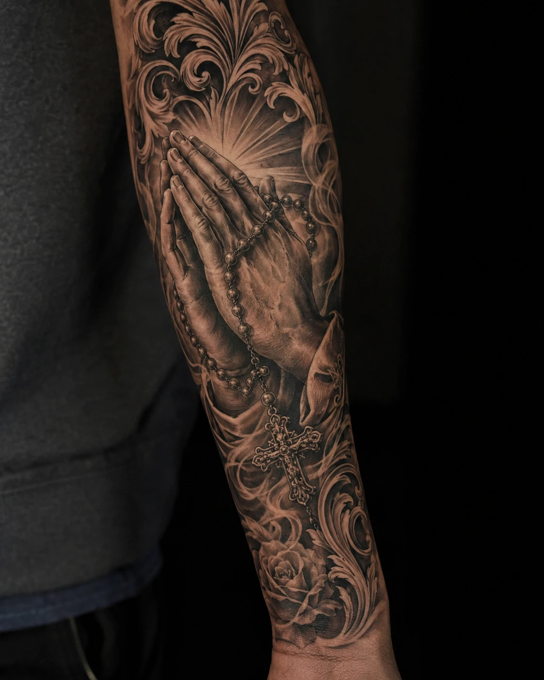 Black and grey forearm tattoo of praying hands, rosary, cross, roses and rays.